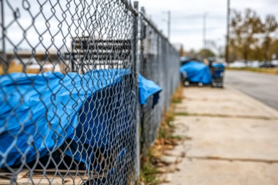 Blue tarps covering tents set up by unhoused people along chain link fence on concrete sidewalk.